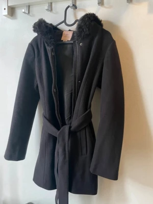 Svart kappa med fuskpäls och huva - Stilren svart kappa från Only med huva kantad av fuskpäls. Kappan har knytband i midjan, dold dragkedja och tryckknappar framtill. Långärmad modell i mjukt material som ger en snygg siluett. Går strax under rumpan på mig som är 162cm. 