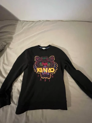 Svart Kenzo tröja med tigertryck - Svart sweatshirt från Kenzo med ikoniskt tigerhuvud i färgglada broderier på bröstet och texten 'KENZO PARIS'. Rund halsringning och långa ärmar. Perfekt statement-plagg för dig som gillar streetwear och snygga detaljer.