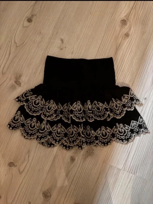 Svart mini volangkjol med broderi - Supersöt svart kortkjol med bred smock i midjan och dubbla volanger. Kjolen har detaljerade vita broderier längs volangerna som ger en unik och trendig look. Perfekt att styla med en enkel topp för en cool outfit.