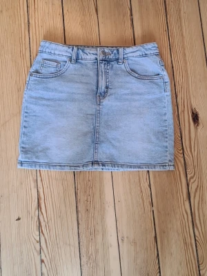 Ljusblå jeanskjol med fickor - Säljer en klassisk ljusblå jeanskjol med fem fickor och knappgylf. Kjolen har en rak passform och är perfekt för dig som gillar en enkel och tidlös stil. Passar till det mesta och är lätt att matcha.