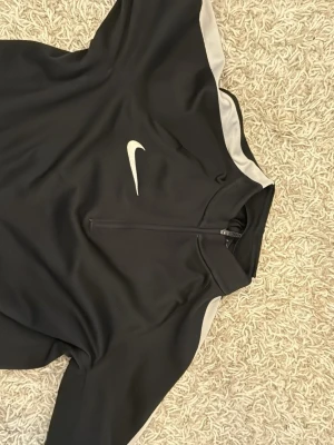 Nike Dri-FIT svart träningströja - Nike Dri-FIT träningströja i svart med vita detaljer och halv dragkedja framtill. Tillverkad i funktionsmaterial som transporterar bort fukt, perfekt för träning och sport. Swoosh-logga på bröstet och kontrastpaneler på axlarna.
