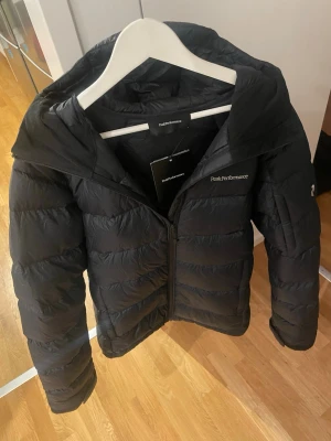 Svart dunjacka från Peak Performance - Säljer en svart dunjacka från Peak Performance med huva och dragkedja framtill. Jackan har en stilren design med diskret logga på bröstet och är fylld med dun för extra värme. Perfekt för kalla vinterdagar och riktigt skön att bära.