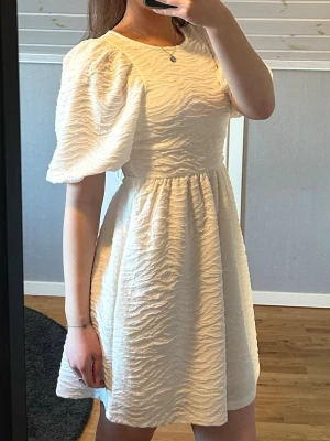 Vit kort klänning med puffärm - Supersöt vit kort klänning med puffärmar och struktur i tyget. Klänningen har öppen rygg med snörning och knyt i ryggen, samt rund halsringning. Perfekt för student eller skolavslutning! Använd fåtal gånger