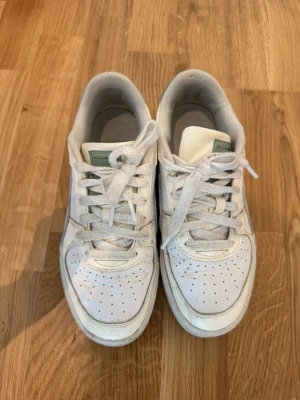 Vita Puma sneakers med grå detaljer - Säljer ett par vita Puma sneakers med grå detaljer på hälen och grönt märke på plösen. Skorna har klassisk låg modell, perforerad tå och robust sula. Materialet är en mix av läder och textil. Perfekta för dig som gillar stilrena och sportiga sneakers.