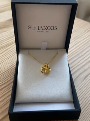 Guldhalsband från Sif Jakobs - Elegant halsband från Sif Jakobs i guldton med ett unikt, flätat hänge. Kedjan är tunn och smidig, och hänget har en modern, organisk form som verkligen sticker ut. Perfekt för dig som gillar stilrena och trendiga accessoarer. Helt ny i orginalförpackning. 