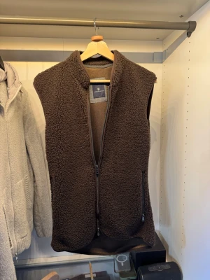 Mörkbrun teddyväst med dragkedja - Mysig mörkbrun teddyväst med hög krage och hel dragkedja framtill. Västen har två fickor med dragkedja och är tillverkad i ett fluffigt, mjukt material. Perfekt för lager på lager och ger en avslappnad, trendig look.