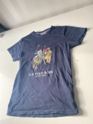 Blå U.S. Polo Assn. t-shirt - Snygg marinblå t-shirt från U.S. Polo Assn. med tryck av två polospelare på hästar och texten 'U.S. POLO ASSN. SINCE 1890' på bröstet. Klassisk passform och rund halsringning. Perfekt till jeans eller shorts.