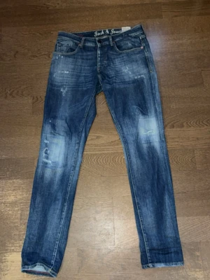 Blå slitna jeans från Jack & Jones - Säljer ett par spicy blå jeans från Jack & Jones med slitningar och ljusare partier på benen. Klassisk femficksmodell med raka ben och låg midja. Perfekta för en avslappnad och trendig stil. 