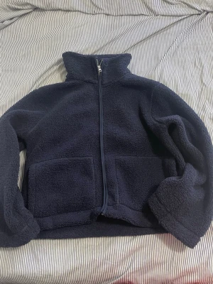 Mörkblå teddyjacka med dragkedja - Mysig mörkblå teddyjacka från lager 157 med hög krage och hel dragkedja framtill. Jackan har två stora fickor på framsidan och är gjord i fluffigt teddy-material som håller dig varm. Perfekt för kyliga dagar och enkel att matcha med olika outfits.