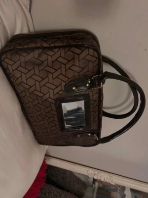 Brun handväska med geometriskt mönster - Snygg brun handväska med svart geometriskt mönster och svarta handtag. Väskan har en rektangulär form och en dekorativ detalj framtill i form av en liten spegel med svart ram .3 innerfack. (Spegeln har små repor)