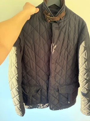 Barbour jacka  - Snygg svart barbour jacka med brun manchesterkrage och detaljer. Jackan har knappar och dragkedja framtill, två stora fickor och en hög krage med spänne. Perfekt för höst och vår när du vill ha en stilren look.