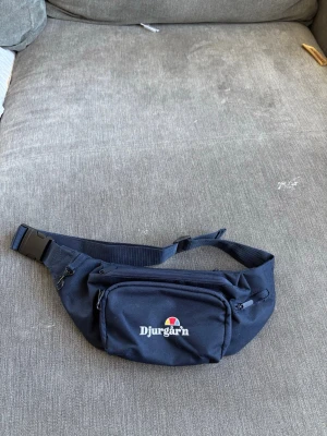 Marinblå magväska Djurgår'n - Snygg marinblå magväska med justerbart band och plastspänne. Väskan har två dragkedjefack och ett mindre ytterfack. Framtill står det 'Djurgår'n' med klubbens färger broderat ovanför texten. Perfekt för att bära det viktigaste på stan eller till matchen.