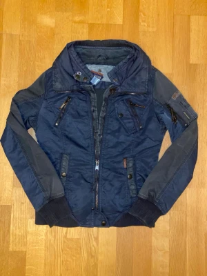 Mörkblå vindjacka från Khujo - Snygg mörkblå vindjacka från Khujo med flera dragkedjeförsedda fickor, hög krage och detaljer i metall. Jackan har ribbade muddar vid ärmslut och nederkant samt patch på ärmen. Perfekt för dig som vill ha en sportig och urban look.