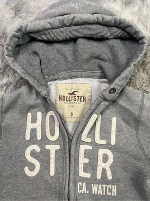 Grå zip-hoodie från Hollister - Grå hoodie från Hollister med dragkedja, stor huva och vita broderade bokstäver på bröstet. Mjuk insida och kängurufickor framtill. Perfekt för chill dagar och enkel att matcha med jeans eller mjukisbyxor. Det är en litet hål på ena ärmen men annars väldigt bra skick.