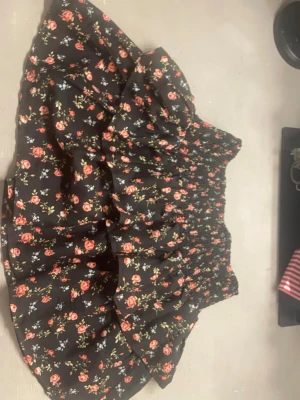 Svart blommig volangkjol - Kort svart kjol med volanger och små blommor i rött, rosa och vitt. Kjolen har bred resår i midjan för skön passform och ett romantiskt blommönster över hela tyget. Perfekt för dig som gillar en söt och trendig stil.