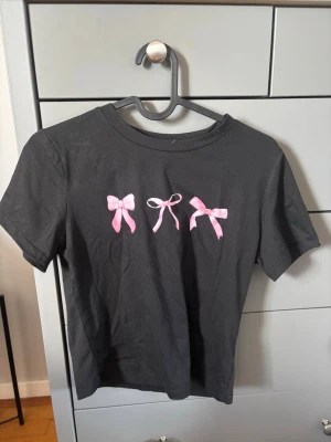 Svart t-shirt med rosa rosetter - Svart t-shirt med tre söta rosa rosetter tryckta på bröstet. Klassisk rund halsringning och korta ärmar. Perfekt för dig som gillar gulliga detaljer och enkel stil.
