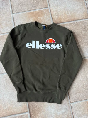 Grön sweatshirt från Ellesse storlek S  - Mörkgrön sweatshirt från Ellesse med stor vit logga och röd-orange detalj på bröstet. Klassisk rund halsringning, ribbade muddar vid ärmslut och nederkant. Perfekt för en avslappnad och sportig stil. Använd en gång, så väldigt fint skick. Nypris 799:- 
