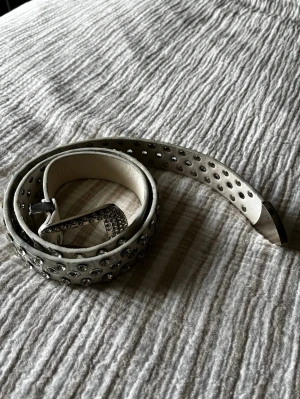 Silvrigt nitarbälte i läder - Säljer ett smalt bälte i silverfärgat läder med massor av små, runda nitar längs hela bandet. Spännet är också dekorerat med nitar för extra edge. Perfekt accessoar för att lyfta vilken outfit som helst.