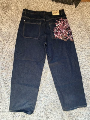 Derschutze Selvedge Indigo Denim Cherry Blossom Brodyr Storlek 34 - Unika mörkblå selvedge indigo denim jeans från Derschutze med bred passform och snygg cherry blossom brodyr på bakfickan och framtill. Jeansen har klassiska fem fickor och är gjorda i Selvedge denimtyg. Perfekta för dig som vill sticka ut med coola detaljer och streetwear-vibe. ALDRIG ANVÄNDA, nyskick! 