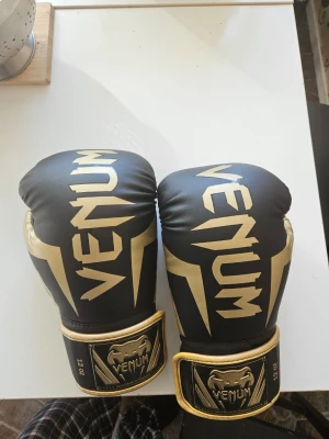 Venum boxningshandskar svart/guld - 🥊 Venum Elite Boxing Gloves – Black/Gold | 12 oz  Selling my Venum Elite boxing gloves in the classic black and gold colorway. Handmade in Thailand, as is standard for Venum's Elite series.  ⚖️ Size: 12 oz 🎨 Color: Black/Gold 🏷️ Brand: Venum Elite 📦 Condition: Used, decent condition – normal wear from use.  Great for boxing, kickboxing or Muay Thai training. Venum Elite is a trusted brand for both beginners and more experienced fighters.  Feel free to message with any questions! 👊