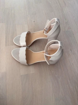 Vita pumps med blockklack från Grace - Säljer ett par stilrena vita pumps från Graceland med bred blockklack och öppen tå. Skorna har en justerbar rem runt ankeln med guldfärgat spänne och en vågig kant framtill. Tillverkade i konstläder med mjuk innersula för extra komfort. Använda några få gånger men har ändå lite små slitningar. Hör av dig vid frågor🤗