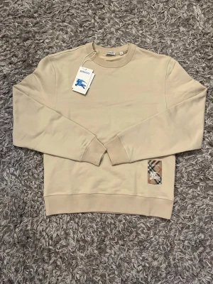 Beige sweatshirt från Burberry - Stilren beige sweatshirt från Burberry med klassisk rund halsringning och ribbade muddar. På framsidan finns en ikonisk Burberry-rutig patch med logga och riddarmotiv. Perfekt för en avslappnad och trendig look. Size m but fits L