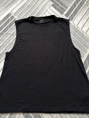 Svart ärmlös linnetopp från Sinsay - En enkel svart ärmlös linnetopp från Sinsay, perfekt för en avslappnad stil. Tillverkad i mjuk bomull med rund halsringning och loose passform. Passar dig som gillar minimalistiska plagg och vill ha något bekvämt till vardags.