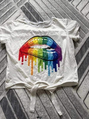 Vit t-shirt med regnbågsläppar och knyt - Snygg vit t-shirt med ett stort tryck av läppar i regnbågens färger på framsidan. T-shirten har korta ärmar och en trendig knytning nedtill. Perfekt för dig som vill sticka ut med färg och coolt motiv.