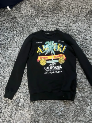 Svart sweatshirt med tryck från Amiri - Cool svart sweatshirt från Amiri med färgglatt tryck framtill som visar palmer, texten 'California', 'Spring Summer' och '2022'. Tröjan har rund hals, långa ärmar och ribbade muddar. Perfekt för dig som gillar streetwear och statementplagg.
