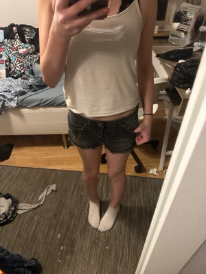 Svarta jeansshorts med fransig kant - Säljer ett par svarta jeansshorts med fransig kant och slitna detaljer. Shortsen har låg midja, klassiska fem fickor och knappgylf. Perfekta för sommaren och ger en cool, avslappnad look.