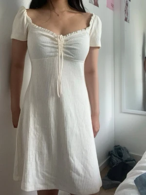 Vit ribbad kortklänning med snörning - Supersöt vit kortklänning med ribbad struktur och snörning framtill. Klänningen har korta puffärmar och en lätt v-ringad urringning. Perfekt för sommarens varma dagar och ger en romantisk vibe.