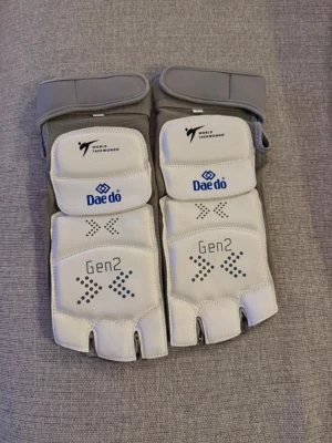 Daedo Gen2 Taekwondo fotsskydd - Säljer ett par vita och gråa Daedo Gen2 E-Foot Protector för taekwondo. Skydden har officiell World Taekwondo-logga och är designade för att ge bra stöd och skydd vid träning och tävling. Materialet är slitstarkt syntetläder med kardborreband för justerbar passform.                     Storlek L  260-265mm