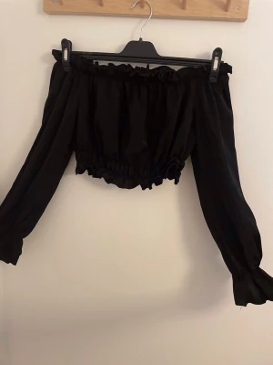 Svart offshoulder croppad topp - Svart offshoulder topp med långa ärmar och croppad passform. Toppen har resår vid axlar och midja för en snygg rynkad effekt och volangdetaljer upptill och nedtill. Perfekt för dig som vill ha en stilren men ändå edgy look.