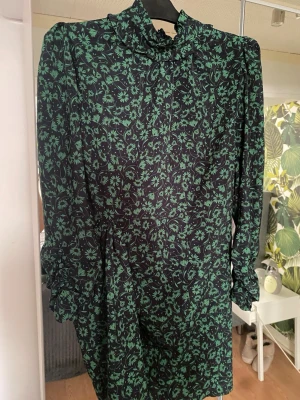 Grönblommig kort klänning från Zara, oanvänd! - Supersnygg kort klänning från Zara med svart botten och grönt blommönster. Klänningen har lång ärm, hög krage med rynk fram och dragkedja i ryggen. Perfekt för dig som gillar trendiga detaljer och vill sticka ut lite extra. Märkt stl XXL men skulle säga att den passar en L.