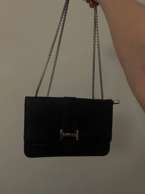 Svart axelväska med kedjerem - Stilren svart axelväska i rektangulär form med silverfärgad kedjerem och metallspänne framtill. Väskan är gjord i ett läderliknande material och har en enkel, modern design som passar till många outfits.