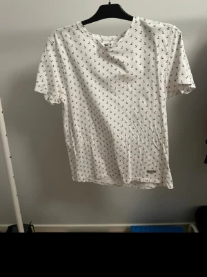 Vit t-shirt med ankarmönster från 157 - Snygg vit t-shirt från 157 med små svarta ankare som mönster över hela tröjan. Klassisk passform med rund hals och korta ärmar. Perfekt för dig som gillar marina vibbar och enkel stil. Tillverkad i mjuk bomull för skön känsla.