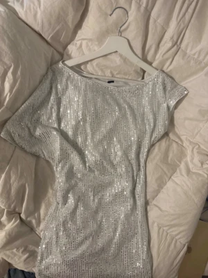 Glittrig silver kortklänning - Säljer en glittrig silverfärgad kortklänning med paljetter. Klänningen har en lös passform, korta ärmar och rund halsringning. Perfekt för dig som vill sticka ut med en riktigt festlig look.