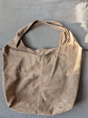 Beige shoppingväska i mocka Becksöndergaard  - En stilren shoppingväska i beige mocka med mjuk känsla och enkel design. Väskan har breda axelband och är rymlig, perfekt för att bära med sig allt man behöver. Snygg och minimalistisk look som passar till många outfits.