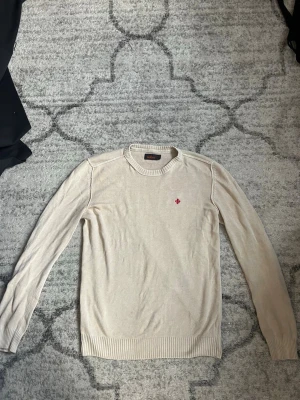 Beige stickad tröja från Woolrich - Snygg beige stickad tröja från Woolrich med rund halsringning och diskret röd broderad logga på bröstet. Tröjan har långa ärmar och ribbade muddar vid ärmslut och nederkant. Perfekt för lager på lager och enkel att matcha.