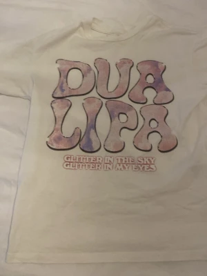 Vit Dua Lipa t-shirt med tryck - Vit t-shirt med stort färgglatt Dua Lipa-tryck på framsidan och texten 'Glitter in the sky, glitter in my eyes'. Klassisk passform och korta ärmar, perfekt för dig som gillar musik och coola prints.