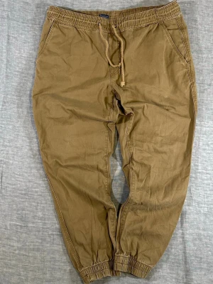 Khaki American Rag Jogger Drawstring Pant Large Tapered fit - American Rag khaki jogger pants with elastic waist and tapered fit. Lightweight cotton, comfortable and easy everyday piece. Clean neutral color that works with anything. Measurements: Waist 46 cm, Rise 27 cm, Thigh 35 cm, Knee 26 cm, Foot 14 cm, Inseam 68.5 cm. Used condition with light wear and minor marks visible in strong lighting, not noticeable in normal use.