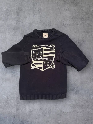 Mörkblå stickad Lexington-tröja - Snygg mörkblå stickad tröja från Lexington med stort vitt emblem och text framtill. Tröjan har rund halsringning, ribbade muddar och är tillverkad i mjukt material. Perfekt för dig som gillar klassisk preppy stil.