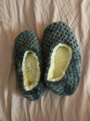 Grå fluffiga tofflor med foder - Mysiga grå tofflor med fluffigt, vitt foder på insidan och mjuk ovansida i strukturmönstrad textil. Sulan är vit med små halkskyddsprickar. Perfekta för att hålla fötterna varma hemma under kalla dagar.