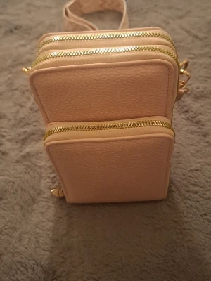 Ljusrosa handväska med guldiga dragkedjor - Säljer en ljusrosa handväska i konstläder med tre stora fack och guldiga dragkedjor. Väskan har en justerbar axelrem och en stilren, fyrkantig form som gör den både praktisk och snygg. Perfekt för dig som vill ha koll på dina grejer och ändå se trendig ut.