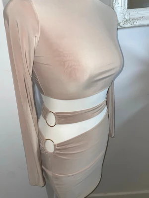 Beige cut out klänning med ringdetaljer - Trendig beige kortklänning med långa ärmar och cut out-detaljer i midjan, dekorerad med två runda ringar i sidorna. Klänningen har en kroppsnära passform och är tillverkad i ett stretchigt, mjukt material som ger en snygg siluett.