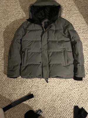 Grå dunjacka från Canada Goose - Säljer en grå dunjacka från Canada Goose i modellen Fusion. Jackan har huva, dragkedja och stora fickor framtill. Perfekt för kalla vinterdagar och riktigt varm tack vare dunfyllningen. Klassisk design med svarta detaljer och diskret logga på ärmen.