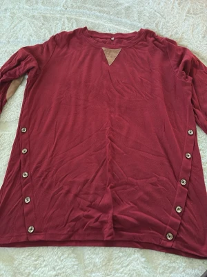 Vinröd långärmad tröja med knappar - Vinröd långärmad tröja med dekorativa knappar längs sidorna och beige detalj vid halsen. Tröjan har även beige armbågslappar som ger en cool kontrast. Mjuk och skön passform, perfekt för dig som gillar en avslappnad stil.