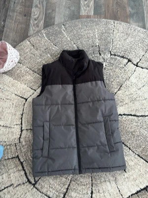 Grå och svart dunväst från primark  - Snygg dunväst i grått och svart från Primark. Västen har hög krage, dragkedja framtill och två fickor på sidorna. Perfekt för lager-på-lager och ger en clean look. Materialet är quiltat och ger en puffig känsla.