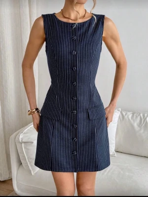 Marinblå randig ärmlös klänning - Snygg marinblå kort klänning med vita vertikala ränder och knappar framtill. Ärmlös modell med rund halsringning och två fickor framtill. Klänningen har en figurnära passform och är tillverkad i ett mjukt tyg, perfekt för en trendig look.