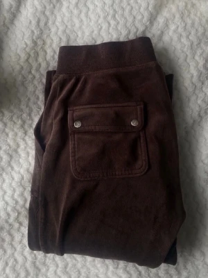 Juicy byxor xs - Mörkbruna mjukisbyxor i mjuk velour med bred resår i midjan och dragsko. Byxorna har stora fickor fram och en bakficka med lock och tryckknappar. Perfekta för chill dagar och riktigt sköna att ha på sig.